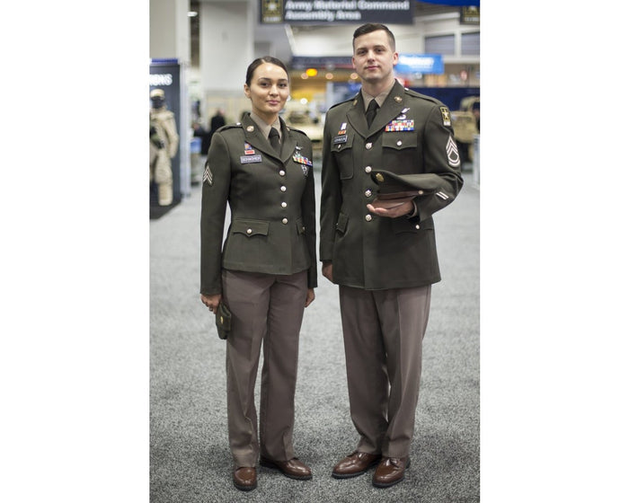 Photograph of two Army sergeants in olive green service uniforms with brown shoes and nametags at an Army Material Command event featuring army vehicles and signage in the background