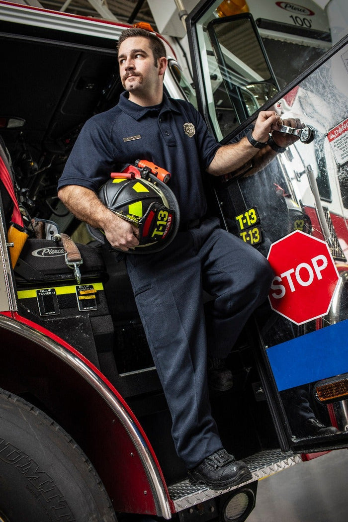 Professional photograph of a firefighter holding a helmet near a red firetruck with T-13 markings, a stop sign, and a Pierce logo visible, showcasing the firefighter's dark uniform, black boots, and bright yellow helmet detail