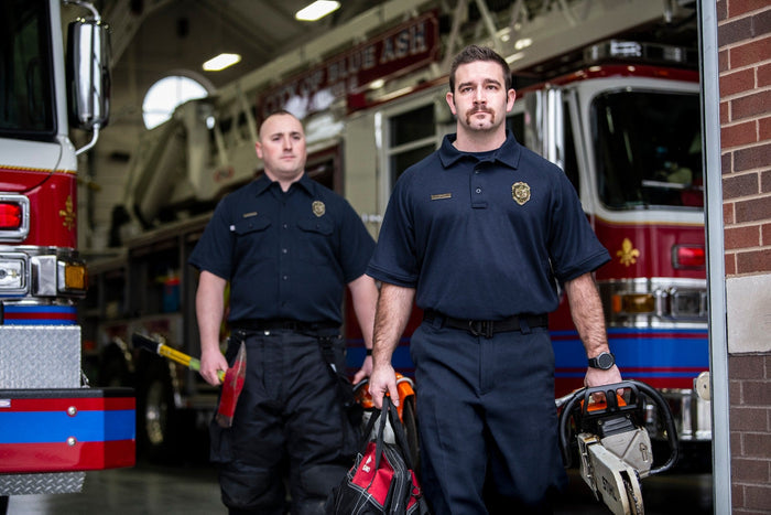 Photograph of two firefighters in a fire station carrying equipment including an axe and chainsaw  in front of a red firetruck with blue and gold details and a fleur-de-lis emblem