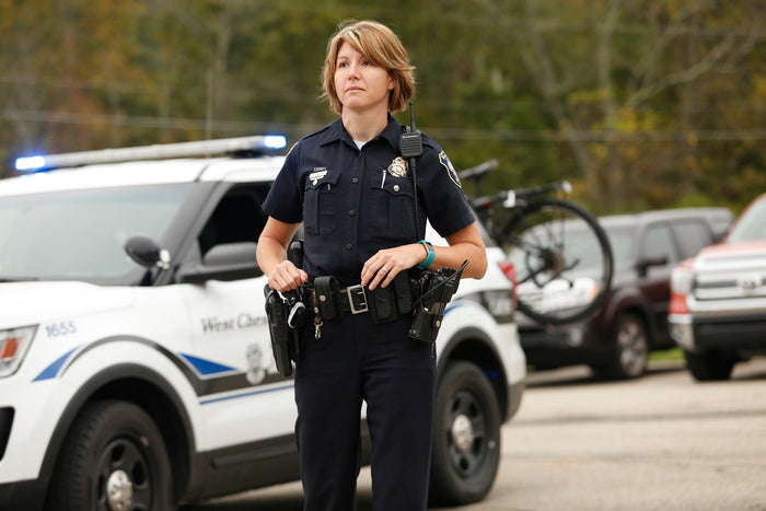 Photograph West Chester Police officer in uniform standing near a police car and bicycle with a teal fitness tracker and a silver badge visible  a  West Chester Police vehicle number 1655 in the background