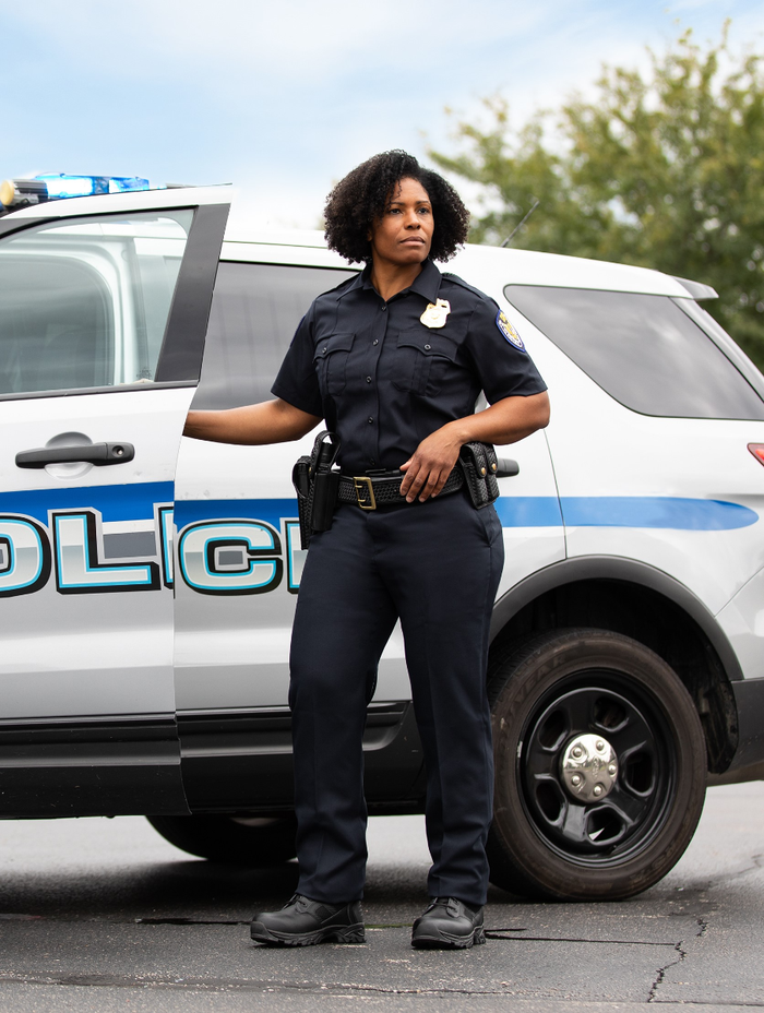 Photograph of a female police officer in uniform standing by a police car with a blue and white design,  in an outdoor setting with leafy trees in the background, wearing black tactical boots and a dark blue shirt with a badge.