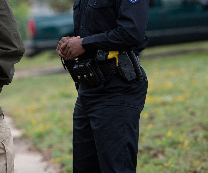 Photograph of a police officer outdoors with a firearm and multiple magazine pouches on their utility belt showing a partially visible dark green vehicle and blurred yellow wildflowers in the background