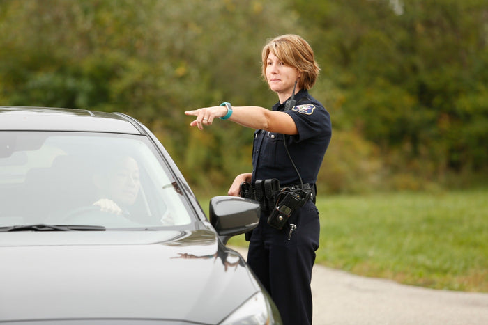 Photograph of a female police officer directing a driver alongside a dark-grey car on a roadside with green trees a teal fitness tracker and a black police radio visible.