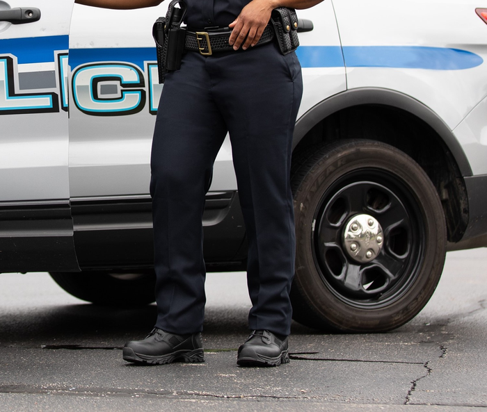 Photograph of a police officer's legs and feet next to a police vehicle with a blue stripe, black boots, a black belt, and a dark asphalt road