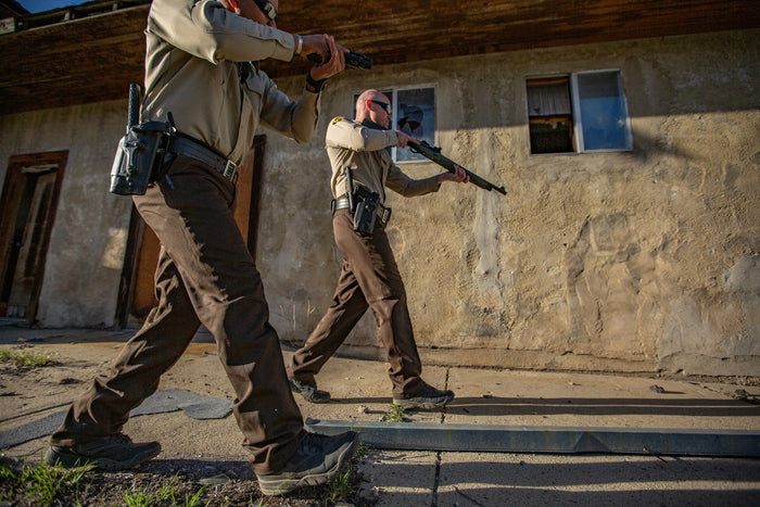 Photograph showing two law enforcement officers in brown uniforms tactical maneuvering near a dilapidated building with a broken window and weathered tan stucco walls carrying handguns and a long gun their dark tactical boots and gear contrasting sharply with the dusty setting