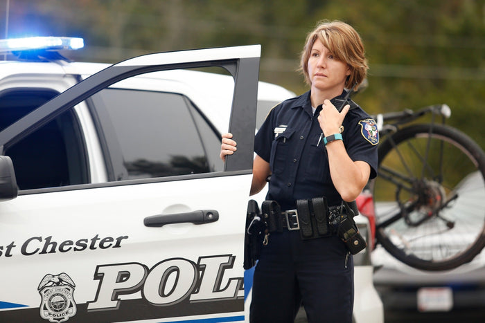 Photograph of a West Chester Police officer using a radio by her police car with a bicycle and flashing blue lights visible in the background showing a teal fitness tracker and a police badge