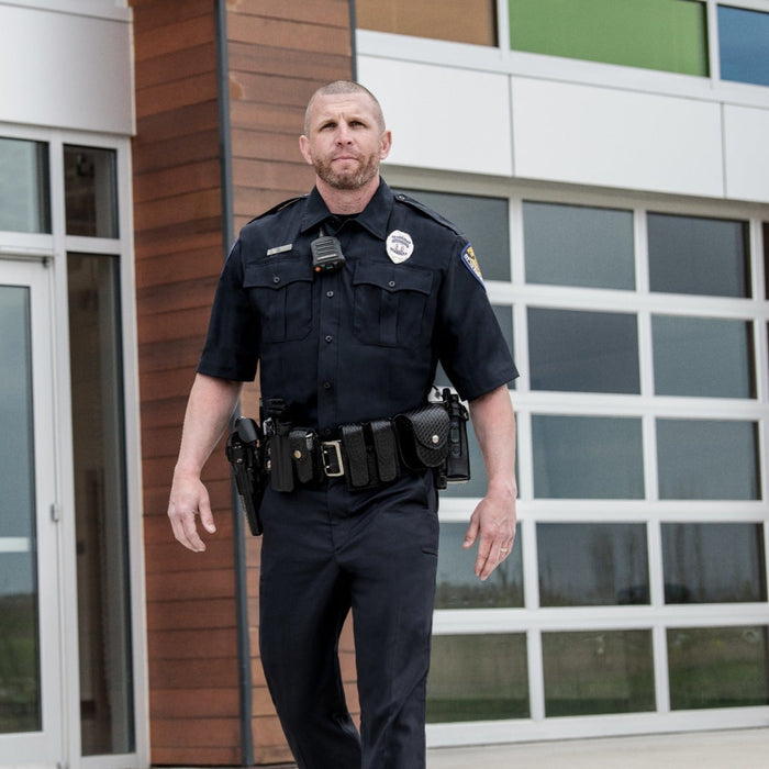 Photograph of a police officer in front of a modern building with large windows and wood paneling,  wearing a dark uniform with a radio and utility belt, showcasing his badge and a serious expression