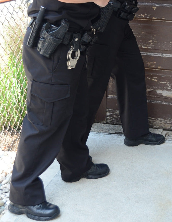 Photograph of two police officers' legs and lower bodies near a weathered wooden door and chain-link fence showing black tactical pants, duty belt with handgun, handcuffs, and shiny black boots.