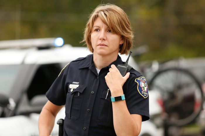 Photograph of a female police officer in West Chester, Ohio, wearing a dark-blue uniform with a Motorola radio, teal fitness tracker, and a West Chester Police patch, standing near a police car with flashing blue lights