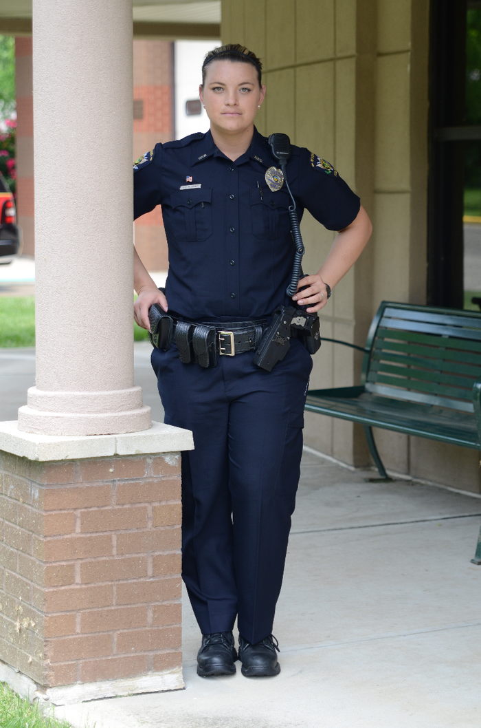 Photograph female police officer in uniform standing near a brick pillar and green bench outside a building with visible tan facade and windows