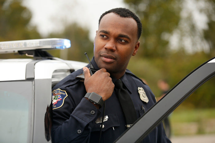 Photograph of a police officer in uniform using a radio next to a police car with flashing lights a dark-colored tie and a smartwatch