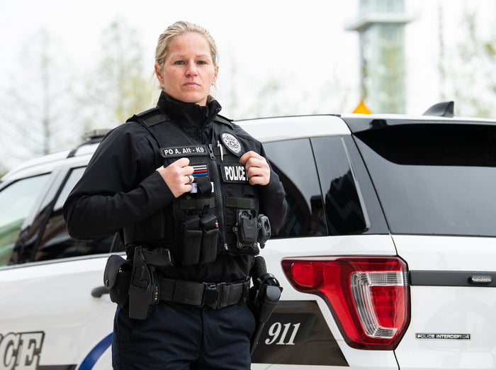 Photograph of a female police officer in uniform with a K9 patch, standing in front of a Police Interceptor SUV with a visible 911 number and red and blue lights, showcasing a thin blue line patch and a police badge.