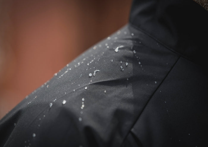 Close-up photograph showing water droplets on a dark gray waterproof jacket's fabric,  with a blurred reddish-brown background suggesting an outdoor setting